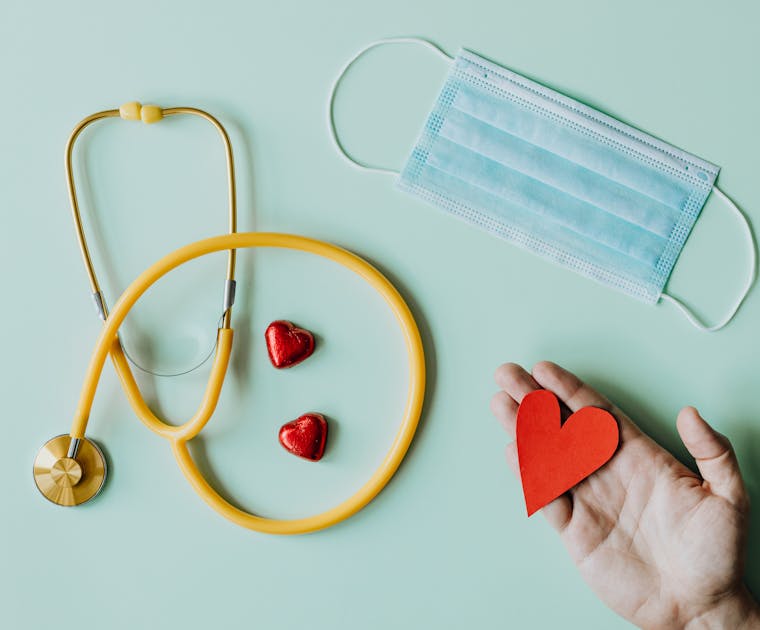 Top view of crop anonymous person hand with red paper heart on table with stethoscope and medical mask for coronavirus prevention