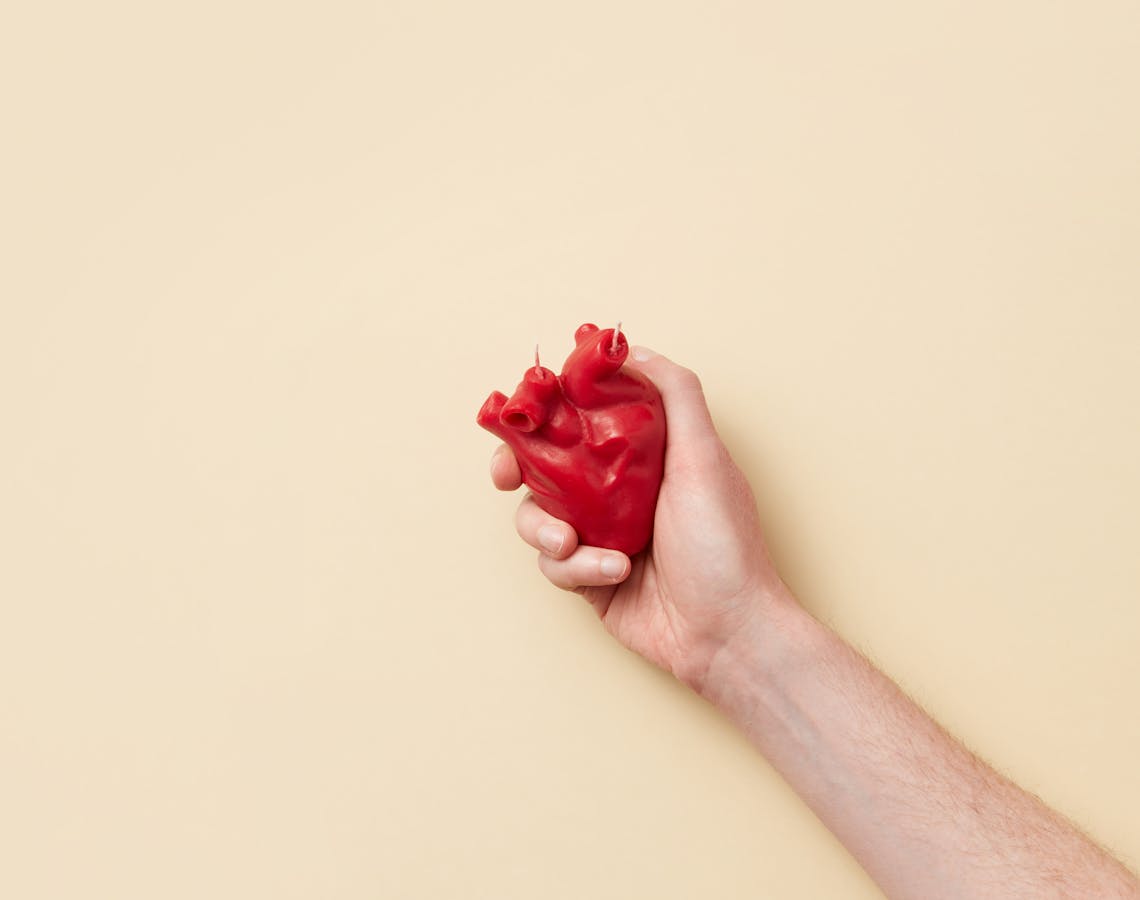 A realistic red anatomical heart model held firmly in a hand against a beige background.