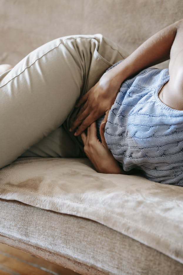 Crop anonymous female in casual outfit writhing on sofa while having stomach ache